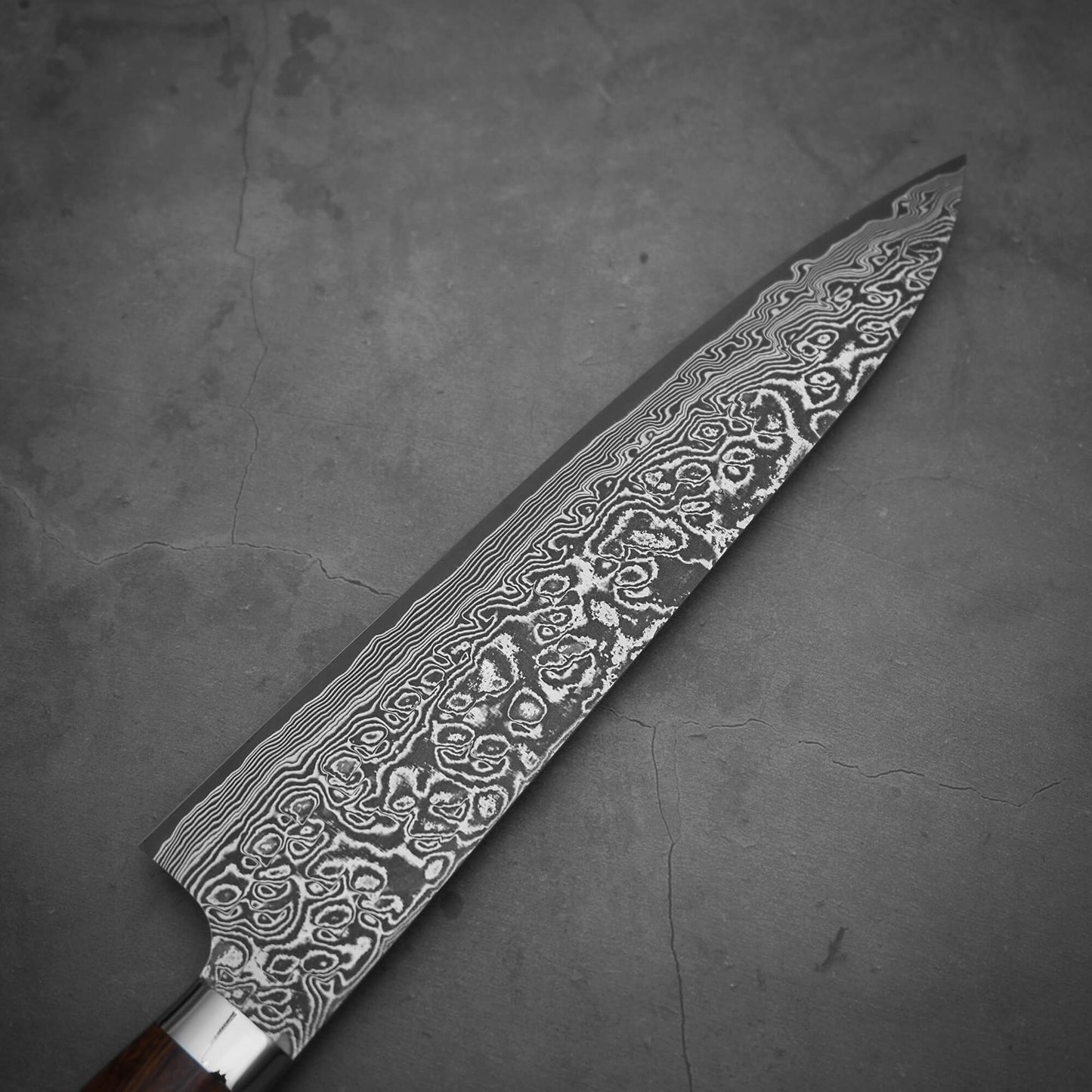 A close-up of the Takeshi Saji black damascus SG2 gyuto ironwood 240mm chef’s knife on a dark grey, cracked surface, highlighting its intricate wavy and swirled patterns on the SG2 stainless steel blade.