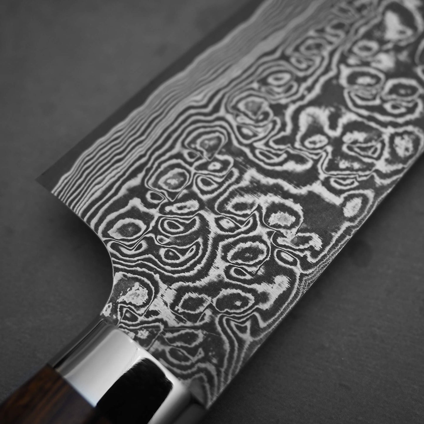Close-up of the Takeshi Saji black damascus SG2 gyuto ironwood 240mm knife, highlighting intricate, wavy patterns on its blade that showcase expert Takeshi Saji craftsmanship. A portion of the handle appears at the bottom left.