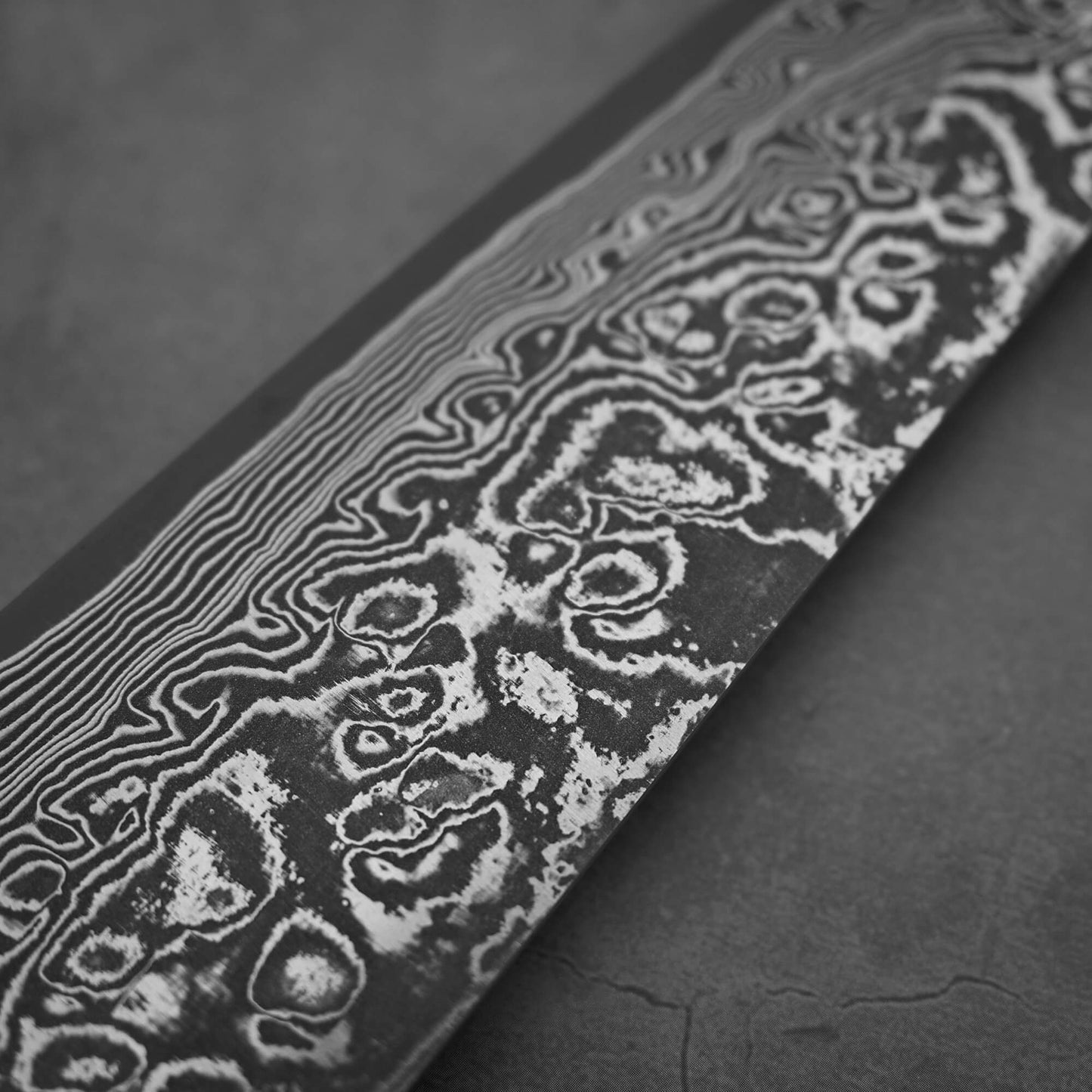 Close-up of the Takeshi Saji black damascus SG2 gyuto ironwood 240mm, featuring wavy patterns on the blade, set against a dark textured background.