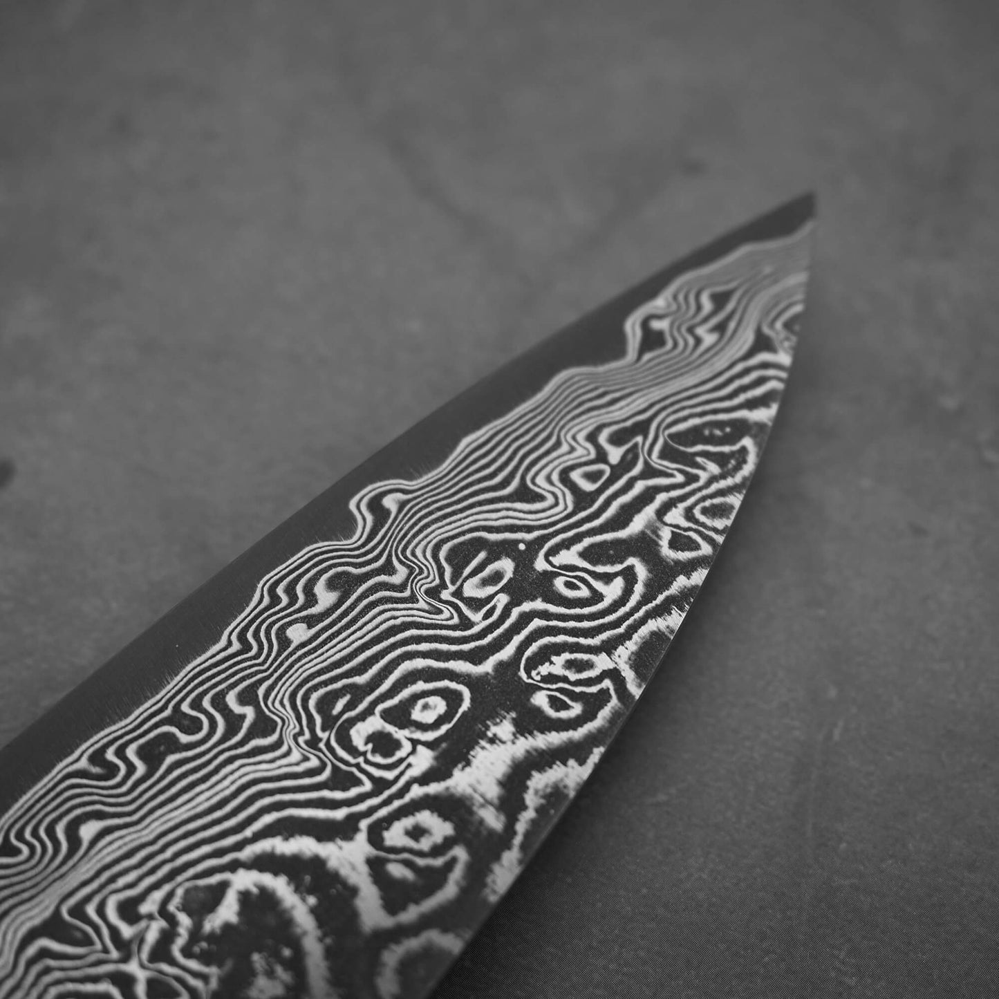 A close-up of the Takeshi Saji black damascus SG2 gyuto ironwood 240mm blade, highlighting its intricate wavy Damascus steel patterns, photographed in black and white on a plain surface.