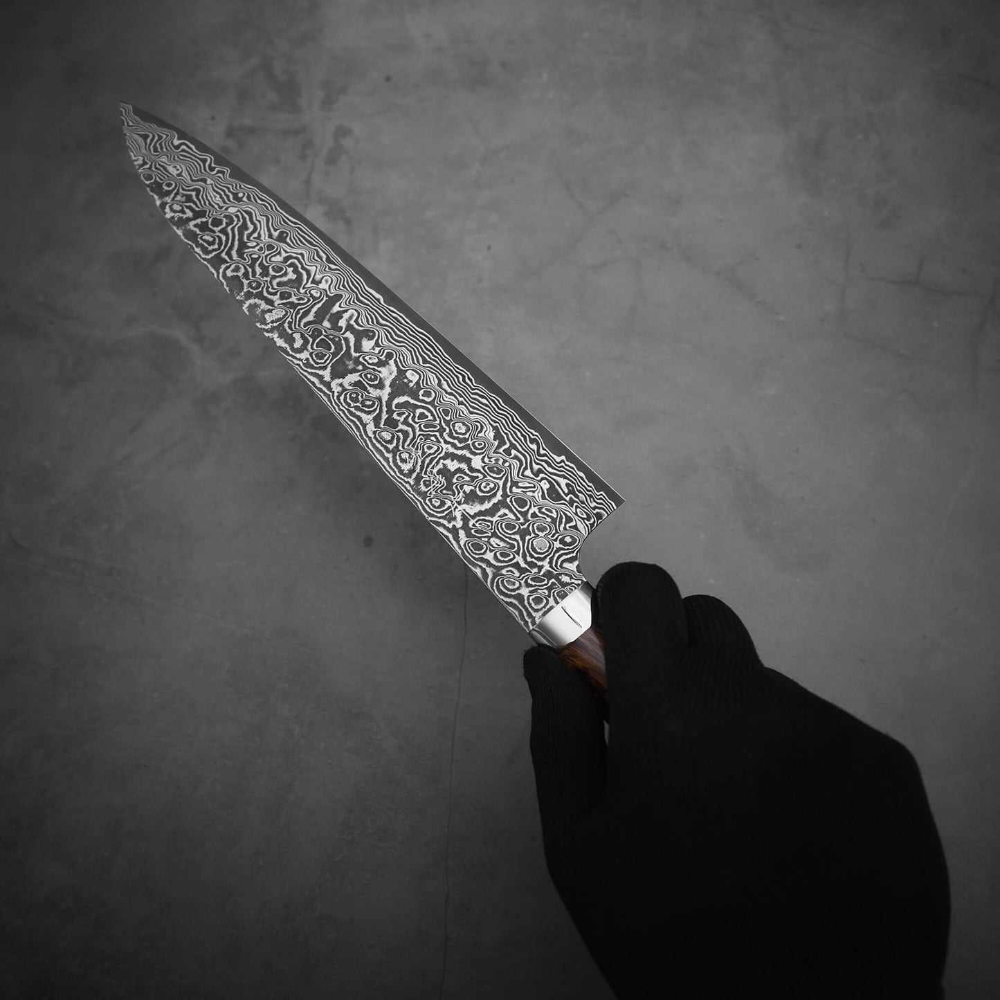 A gloved hand holds a Takeshi Saji black damascus SG2 gyuto ironwood 240mm chef’s knife with a patterned stainless steel blade and ironwood handle, set against a plain gray background.