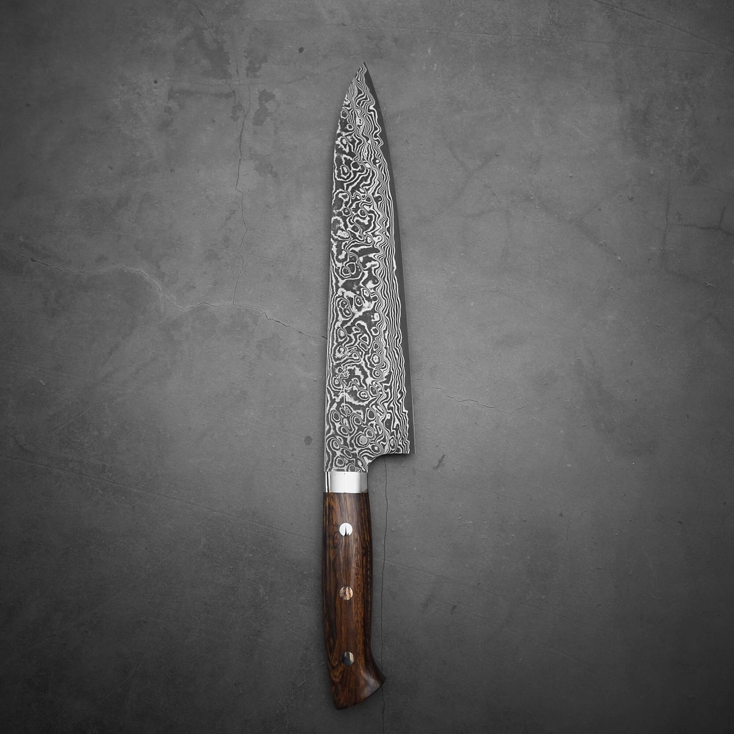 A Takeshi Saji black damascus SG2 gyuto ironwood 240mm chef’s knife with a patterned blade and ironwood handle rests on a dark gray textured surface.