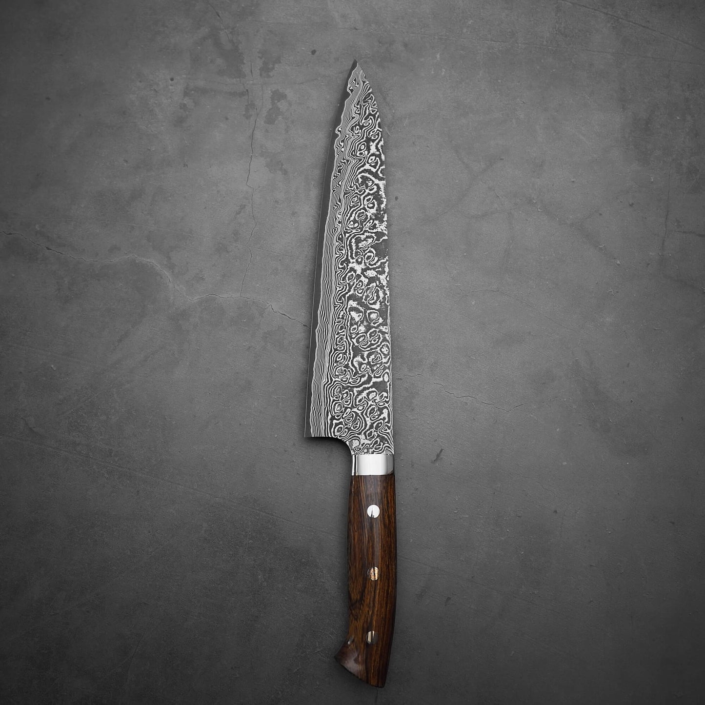 A Takeshi Saji black damascus SG2 gyuto 240mm with an etched blade and ironwood handle featuring three metal rivets rests on a dark, textured surface.