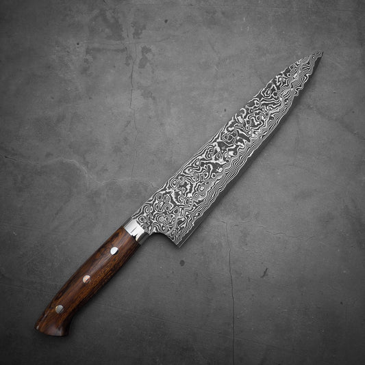 The Takeshi Saji black damascus SG2 gyuto ironwood 240mm, a large black patterned blade with a brown ironwood handle, rests on a gray textured surface.