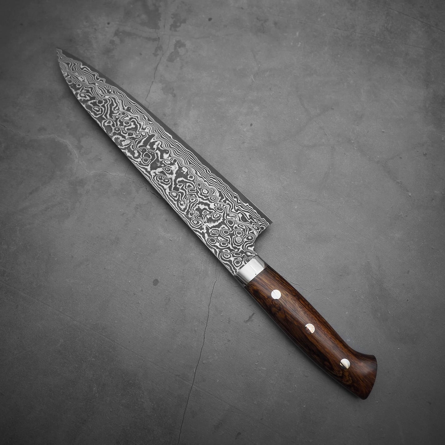 A Takeshi Saji black damascus SG2 gyuto ironwood 240mm chef’s knife with a patterned blade and wooden handle rests on a gray textured surface.