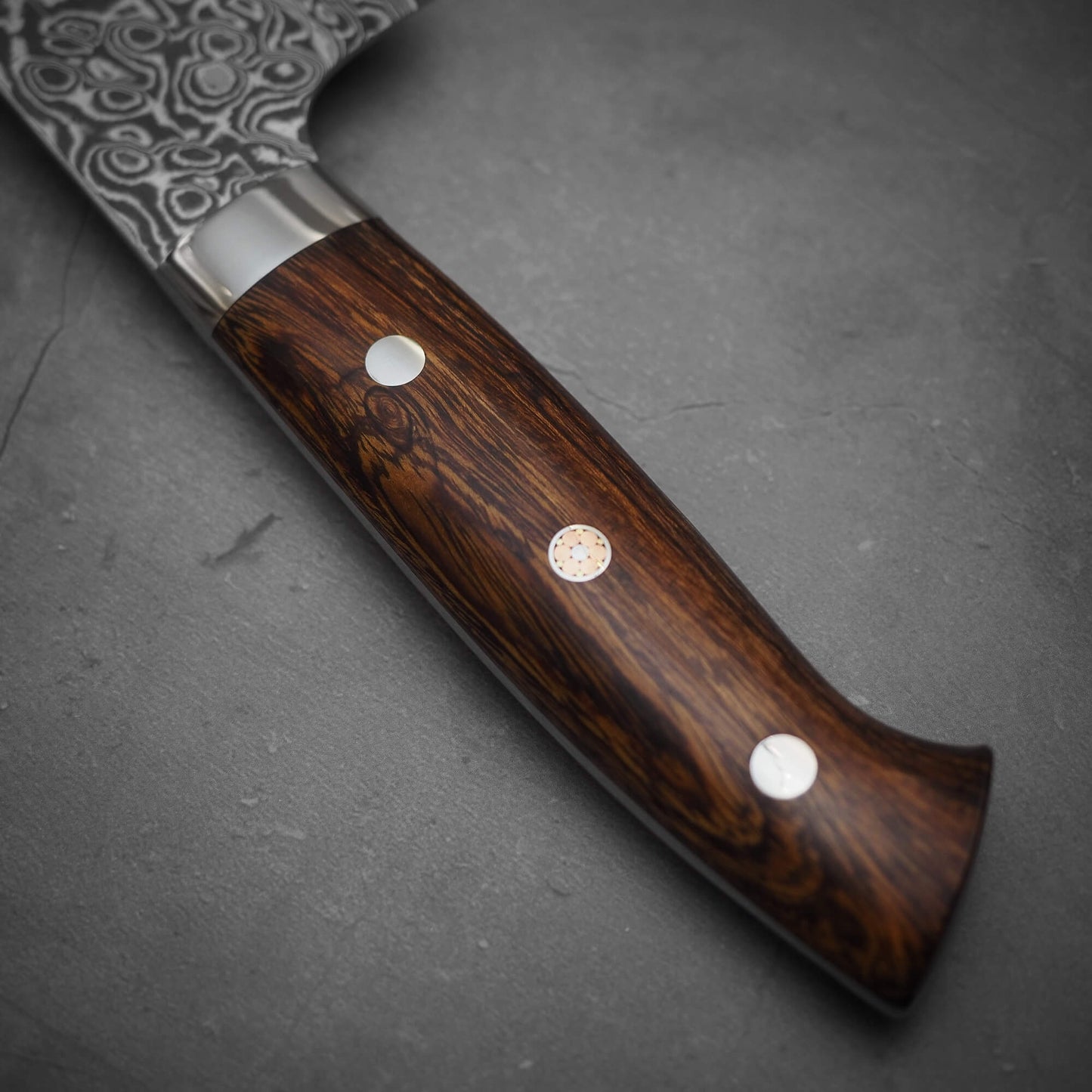 Close-up of a Takeshi Saji black damascus SG2 gyuto ironwood 240mm kitchen knife with a polished dark wood handle and three rivets. The patterned stainless steel blade rests on a gray surface.