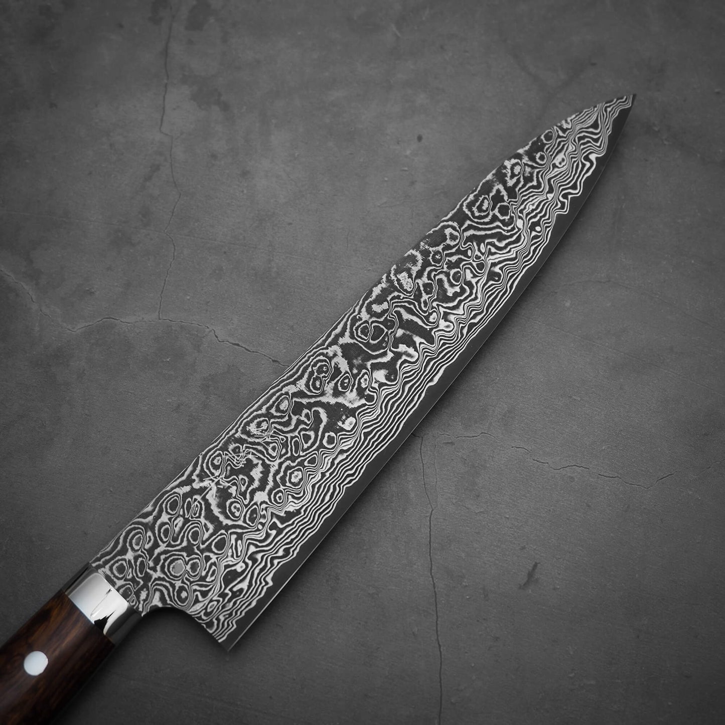 A close-up of the Takeshi Saji black damascus SG2 gyuto ironwood 240mm, showcasing its patterned SG2 stainless steel blade and ironwood handle, resting on a dark textured surface.