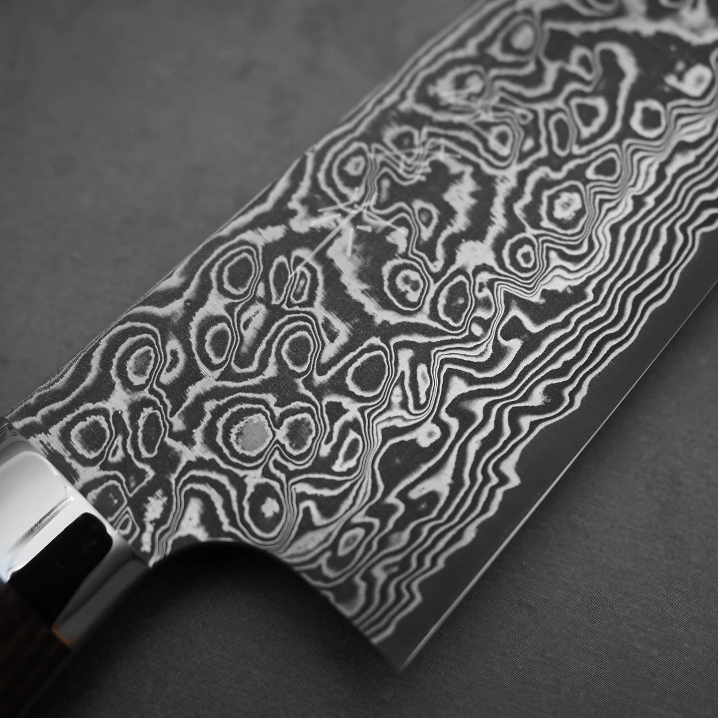 Close-up of the Takeshi Saji black damascus SG2 gyuto ironwood 240mm blade, highlighting intricate steel patterns. The ironwood handle appears partially on the left, all set against a dark, smooth background.