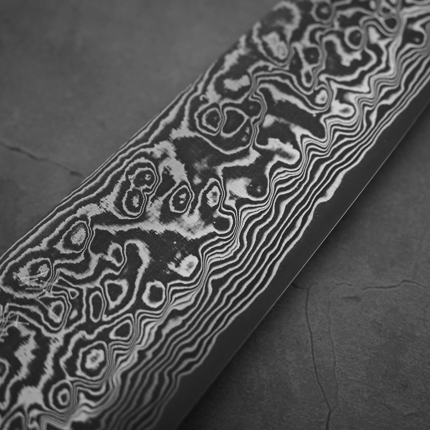 Close-up of the Takeshi Saji black damascus SG2 gyuto ironwood 240mm shows intricate, wavy layers in the SG2 stainless steel blade by Takeshi Saji, with striking swirls set against a dark, textured background.