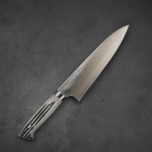 The Takeshi Saji rainbow damascus aogami#2 gyuto 210mm with stag handle lies on a dark, textured surface. Its patterned blade reflects light and is angled slightly to the right.