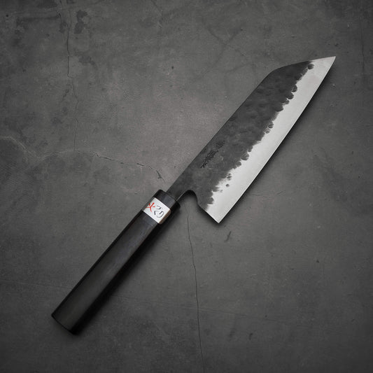 A Teruyasu Fujiwara Denka ebony santoku 180mm knife with a hammered blade and dark wooden handle rests on gray cracked concrete. A small label with red and black text is near the handle. Brand: Teruyasu Fujiwara.