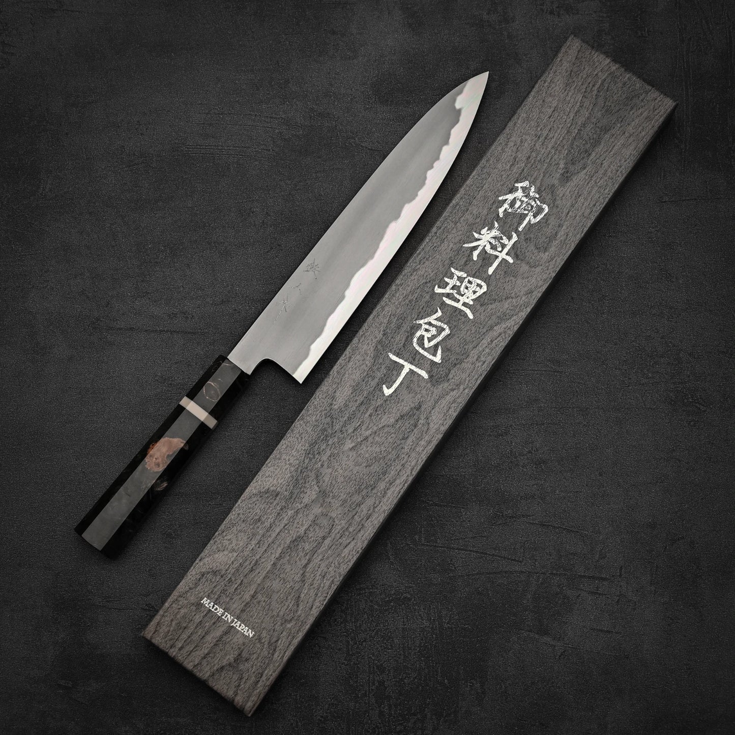 A Tetsujin aogami#2 gyuto 240mm chefs knife with a patterned blade and dark handle rests next to its branded wooden box with white Japanese characters, both displayed on a dark textured surface.