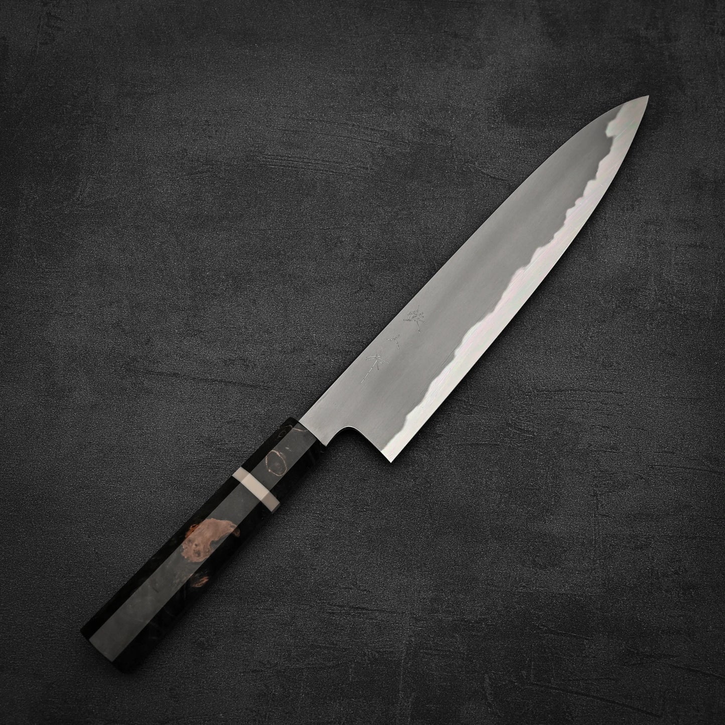 The Tetsujin aogami#2 gyuto 240mm Japanese chefs knife by Tetsujin, featuring a patterned steel blade and dark textured handle, rests on a dark stone-like surface.