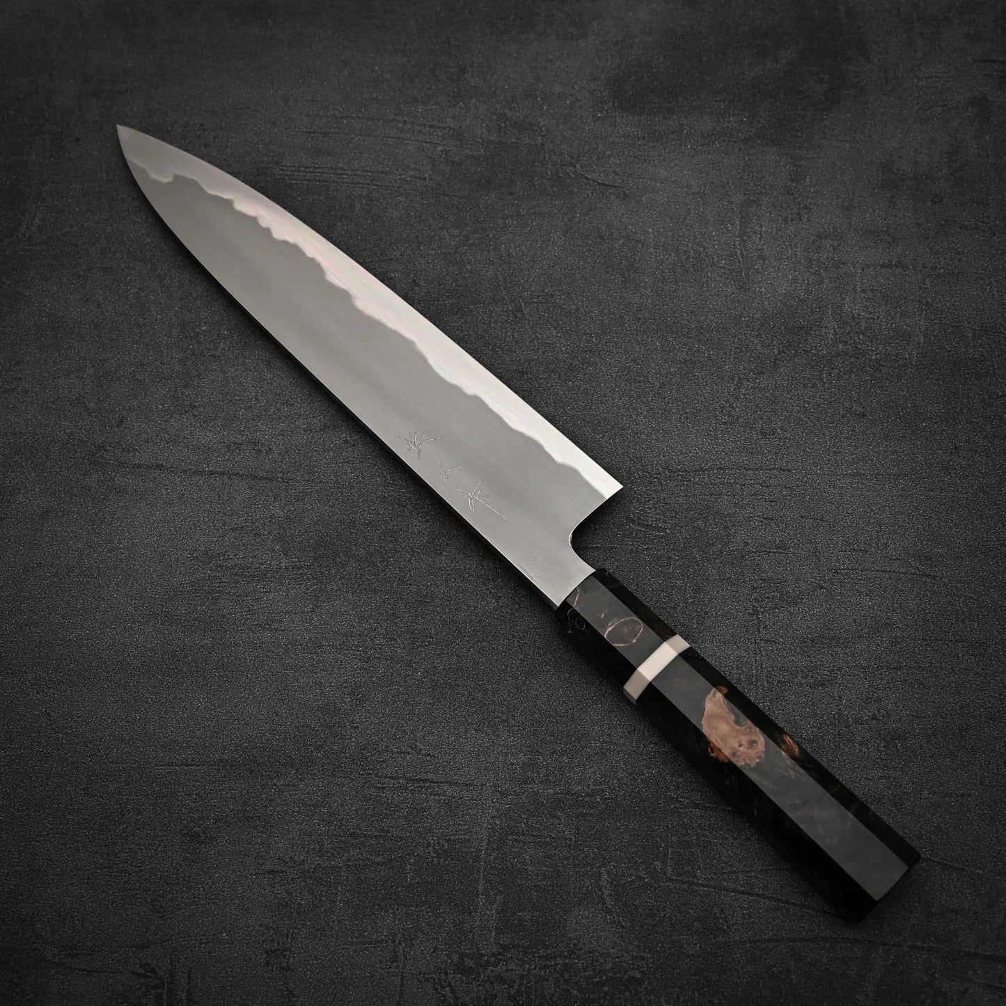 A Tetsujin aogami#2 gyuto 240mm Japanese chefs knife with a polished blade and dark octagonal handle lies diagonally on a textured dark gray surface.