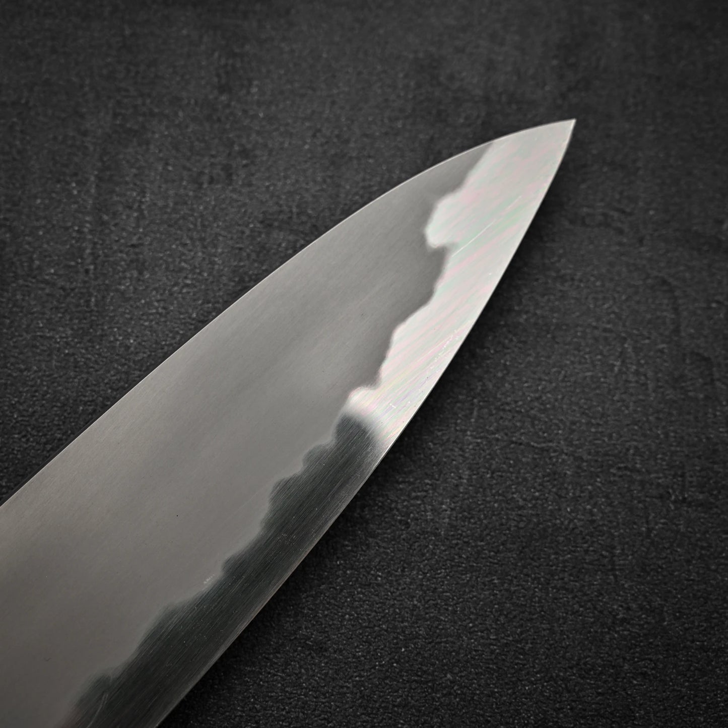 A close-up of the Tetsujin aogami#2 gyuto 240mm chefs knife tip shows its polished blade and wavy hamon pattern set against a dark, textured background.