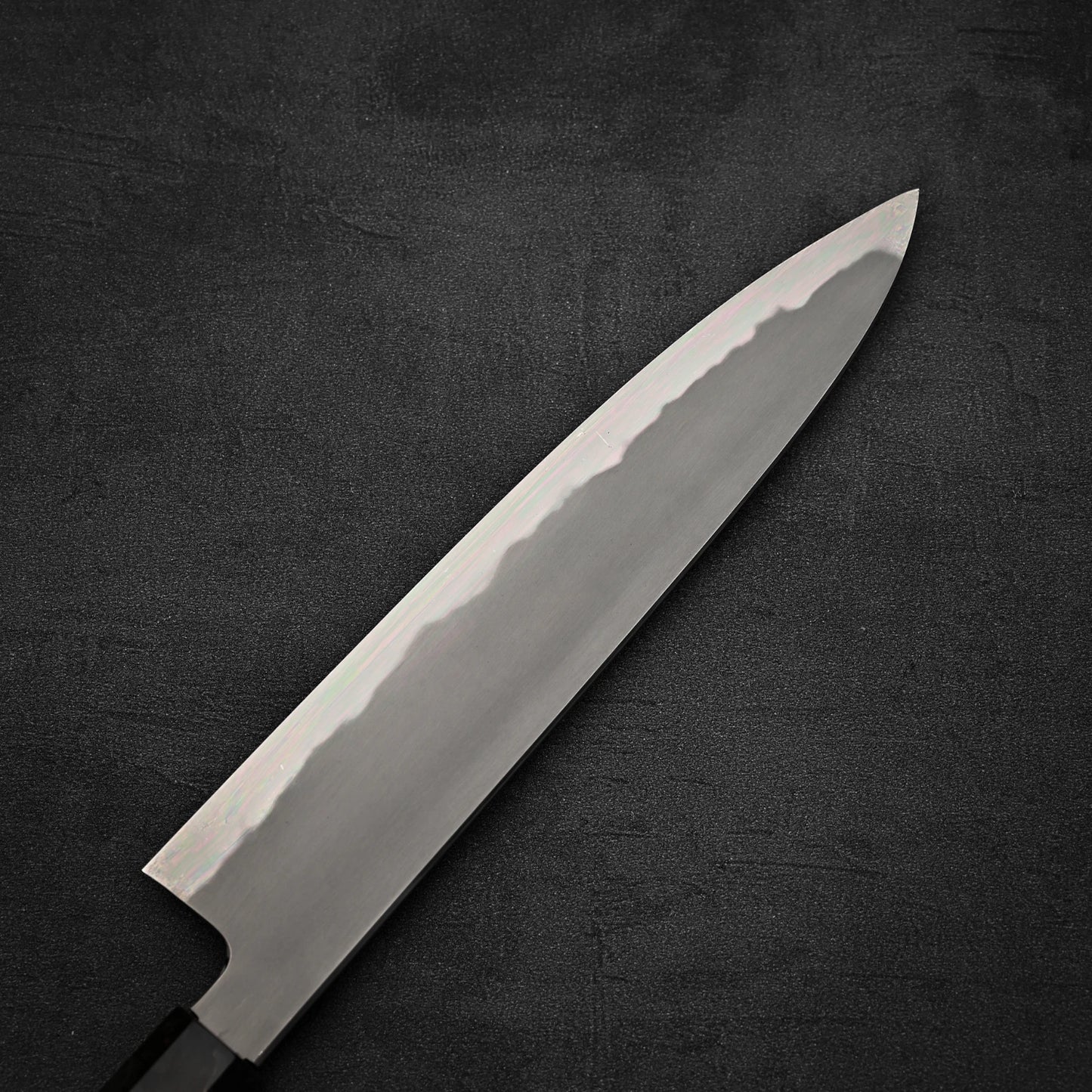 A close-up of the Tetsujin aogami#2 gyuto 240mm by Tetsujin, showcasing its polished blade, sharp edge, and distinctive wavy pattern, set against a dark textured background.