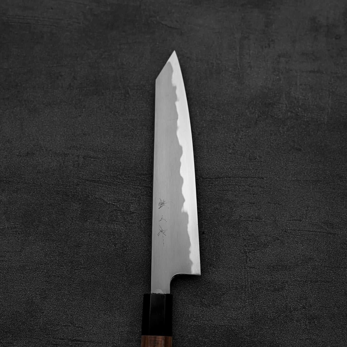 The Tetsujin kasumi aogami#2 petty knife 165mm, featuring a sharp pointed blade and wooden handle, rests on a dark textured surface. Subtle engravings and the kasumi finish by Tetsujin are visible near the base of the blade.