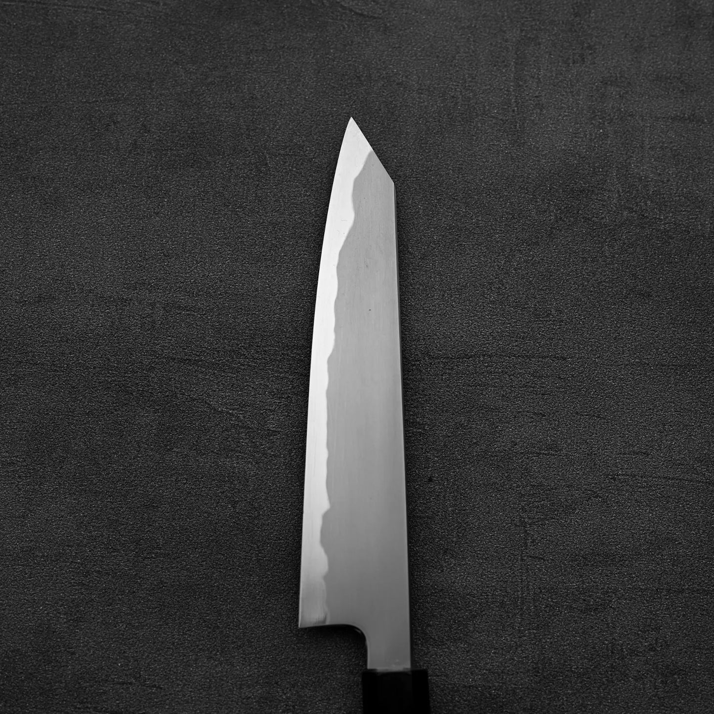 The Tetsujin kasumi aogami#2 petty knife 165mm by Tetsujin, featuring a sharp, pointed blade and kasumi finish, rests on a dark textured surface in an overhead shot.