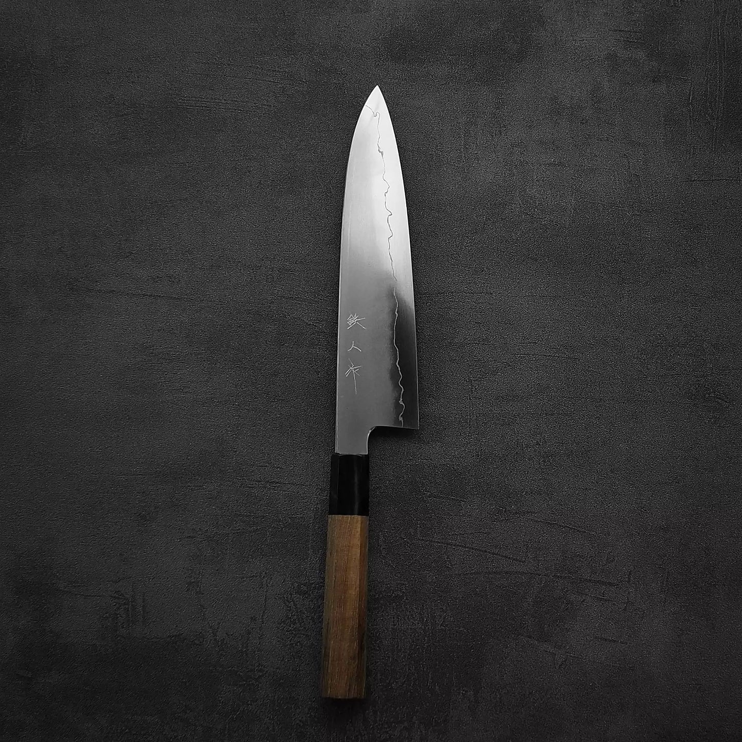 A Tetsujin kasumi ginsan gyuto 210mm chefs knife by Tetsujin, featuring a wooden handle and a silver blade with Japanese engravings, rests flat on a dark, textured background.