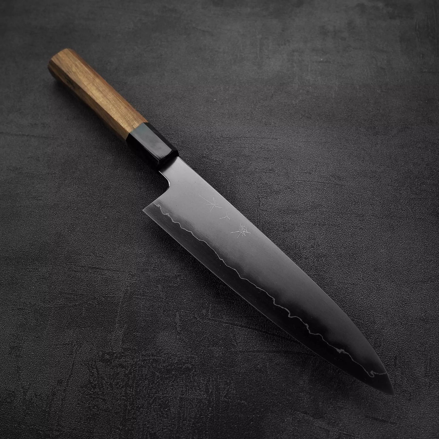A Tetsujin kasumi ginsan gyuto 210mm chefs knife by Tetsujin, with a dark blade, wooden handle, subtle engraved markings, and a wavy pattern near the edge, rests on a textured dark surface.