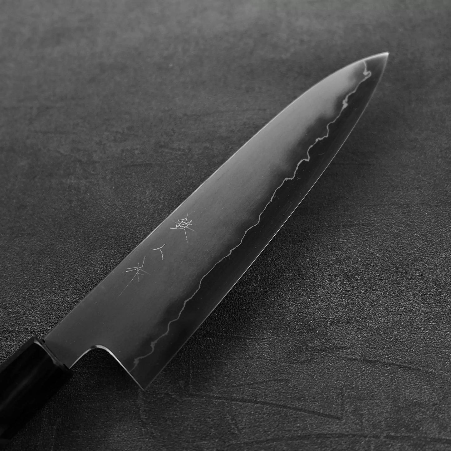 The Tetsujin kasumi ginsan gyuto 210mm by Tetsujin showcases a polished, wavy-pattern blade with Japanese engravings, elegantly placed on a dark textured surface.