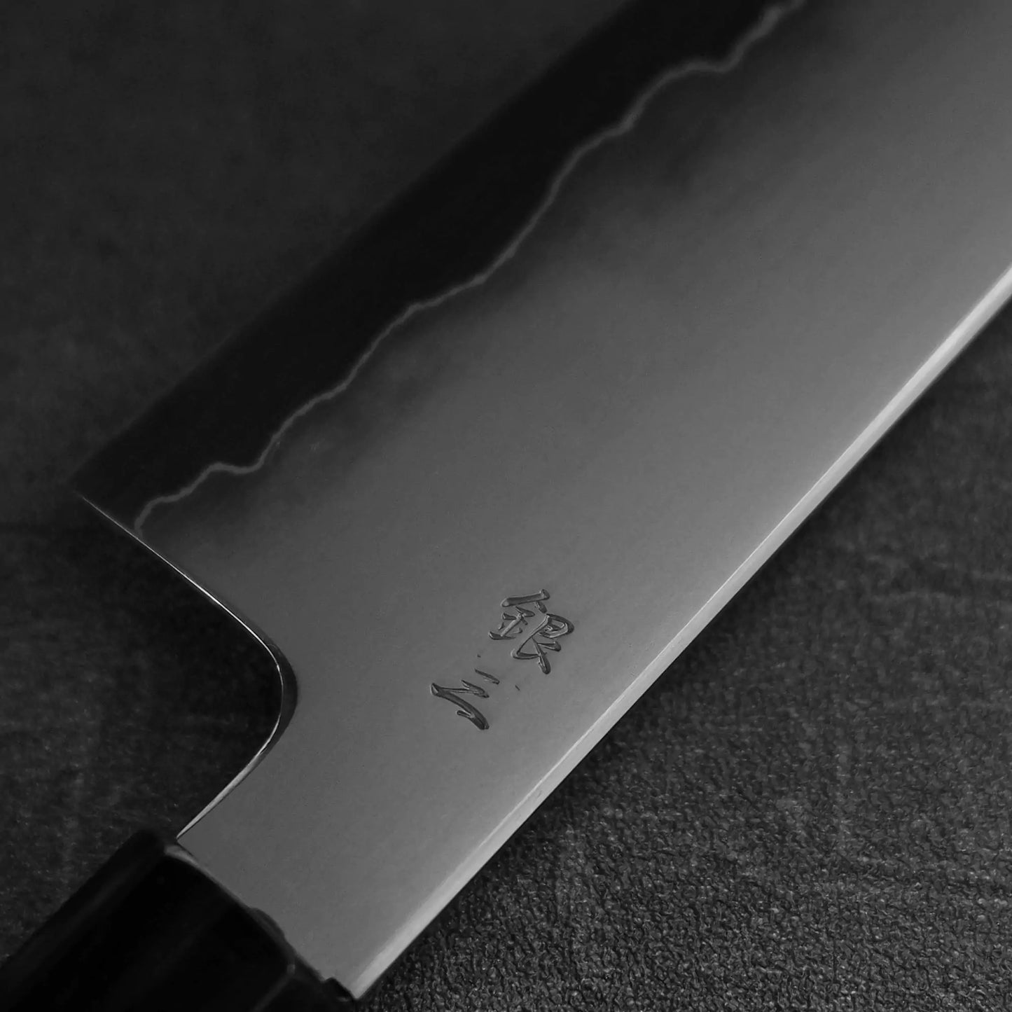A close-up of the Tetsujin kasumi ginsan gyuto 210mm by Tetsujin—a high-performance chef’s knife with a polished, sharp blade, visible hamon line, and engraved kanji—resting on a dark textured surface.