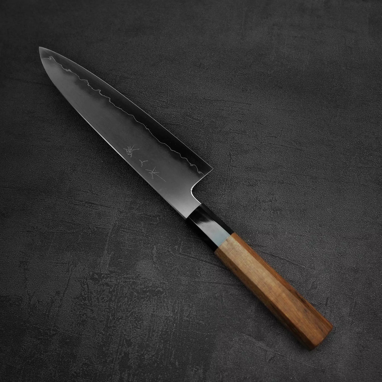 The Tetsujin kasumi ginsan gyuto 210mm by Tetsujin features a patterned dark steel blade with engraved characters and a wooden handle, resting on a textured dark surface—a high-performance chefs knife crafted for culinary precision.
