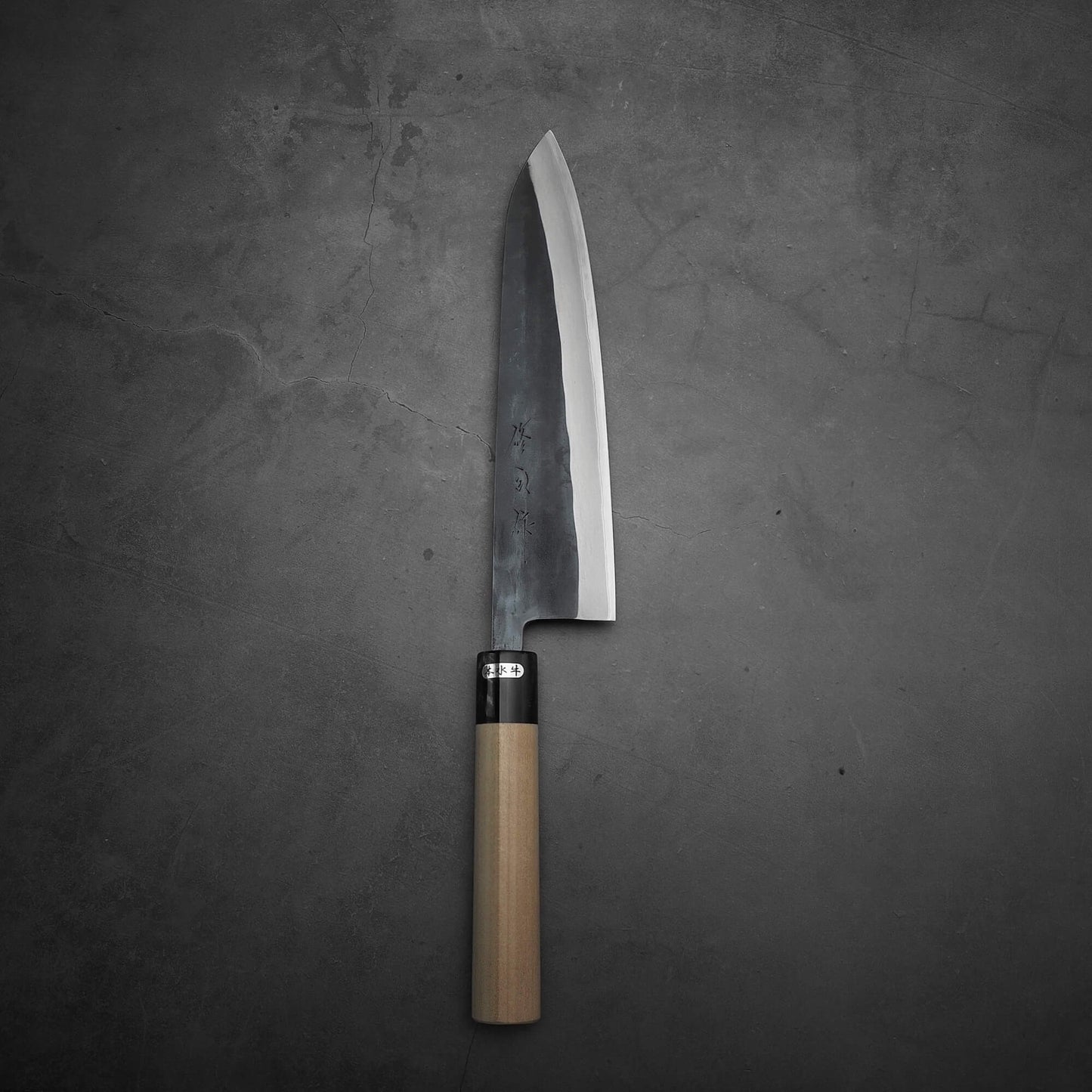 The Toyama kurouchi aogami#2 240mm gyuto, with a dark wide blade and light wooden handle, from Toyama, is forged from aogami #2 steel and rests on a gray textured surface.