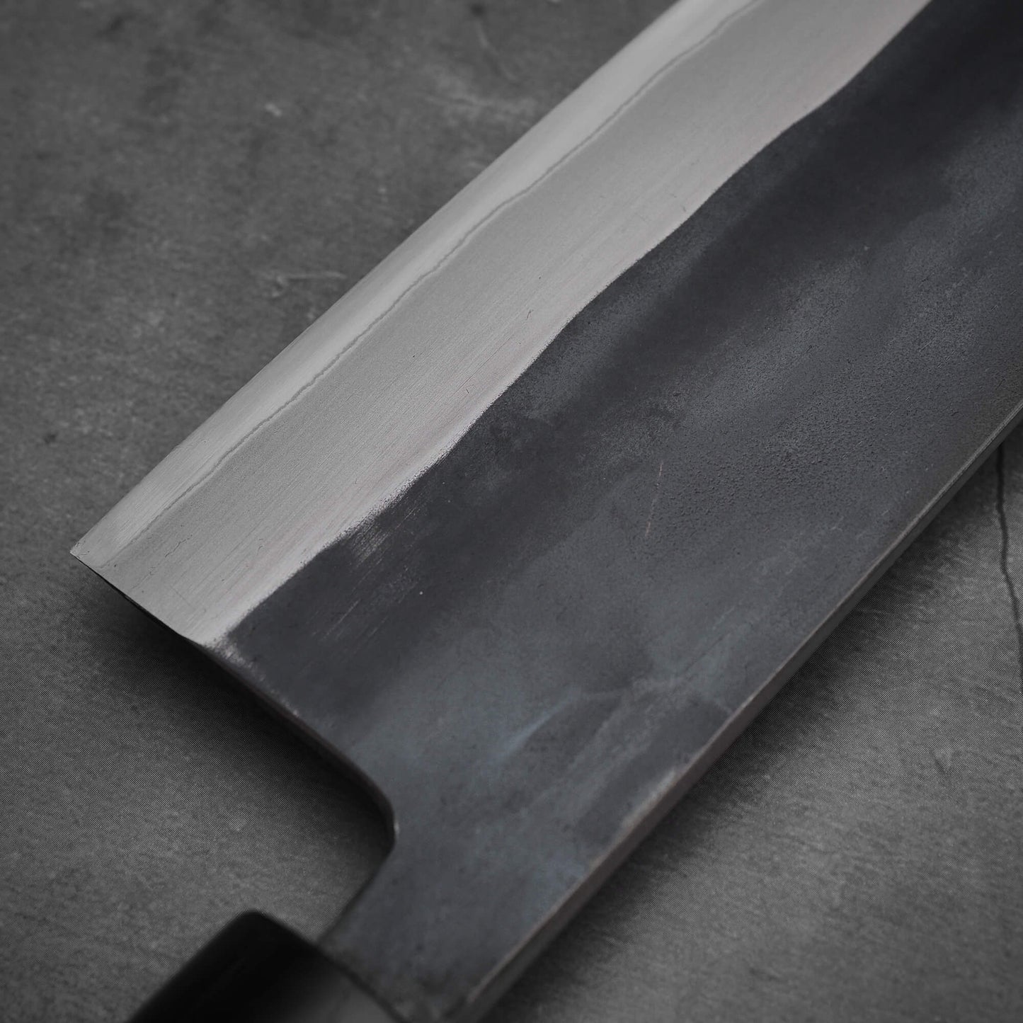 A close-up of the Toyama kurouchi aogami#2 240mm gyuto knife blade by Toyama, displaying a visible hamon line, matte finish, and straight edge against a dark gray stone background, showcasing fine craftsmanship in aogami #2 steel.