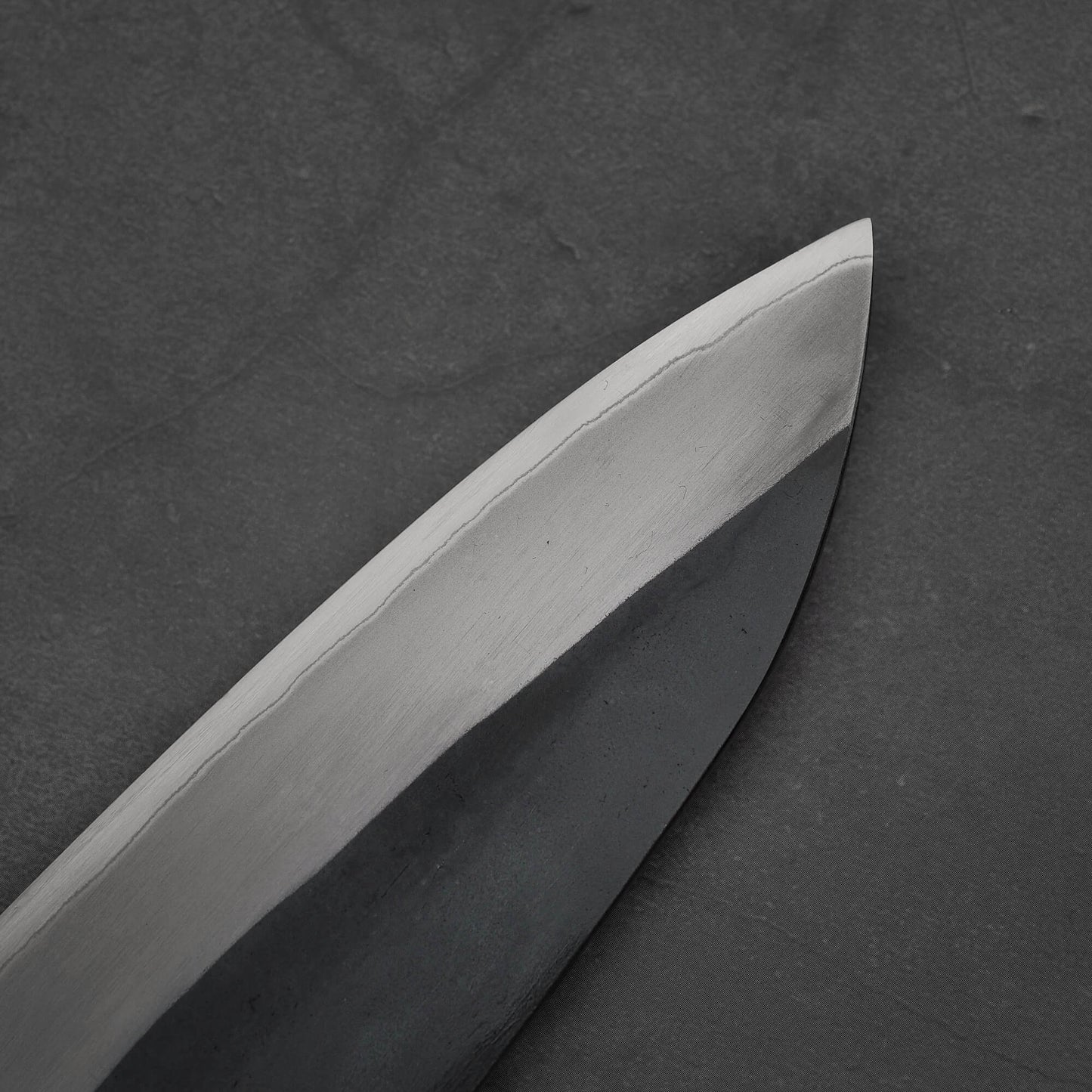 Close-up of the Toyama kurouchi aogami#2 240mm gyuto, highlighting its pointed tip, sharp edge, and contrasting metal tones with a kurouchi finish against a dark gray textured background.