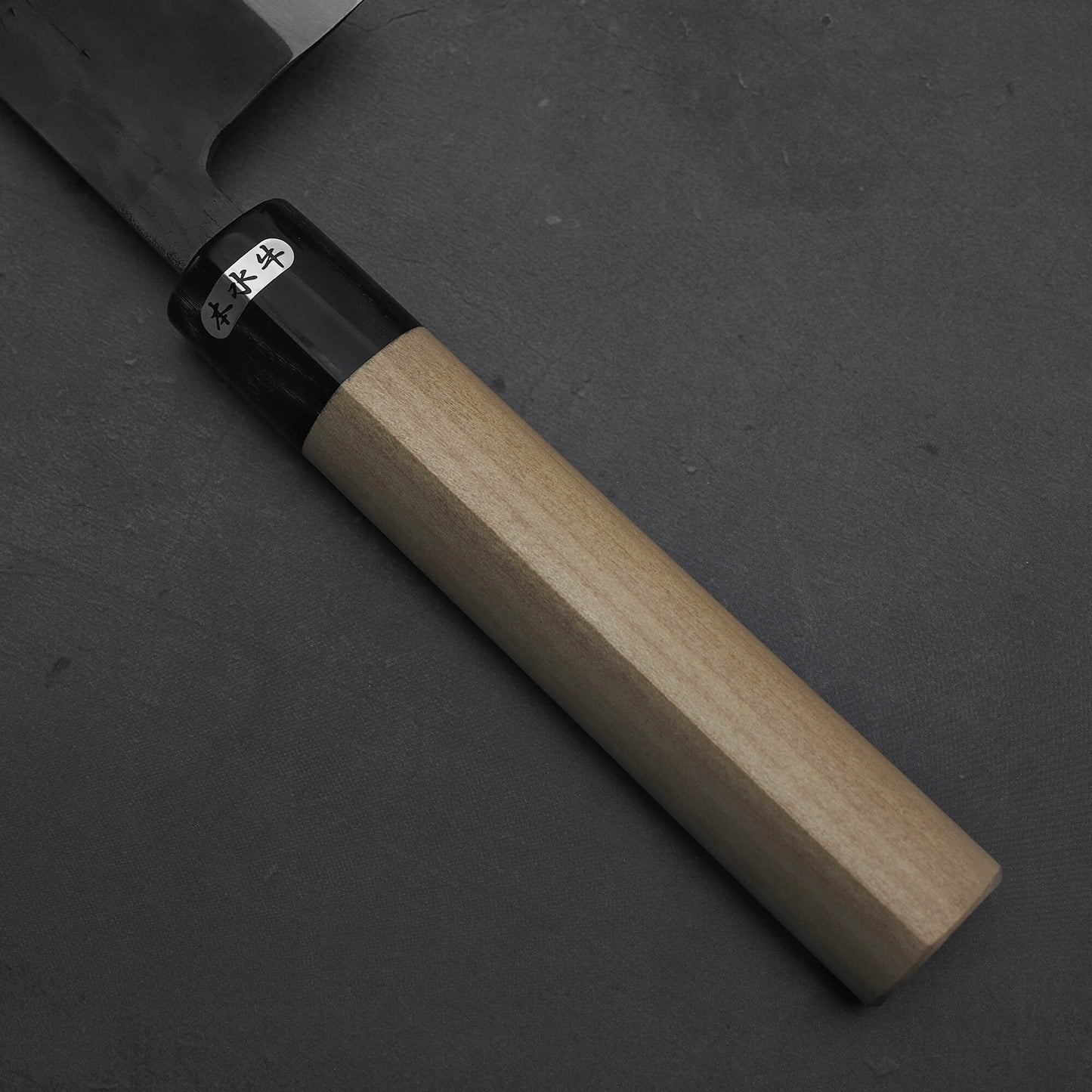 A close-up of the Toyama kurouchi aogami#2 240mm gyuto shows its light wooden handle, black collar, and stainless steel blade with a small white label bearing Japanese characters near the collar, set against a dark gray background.