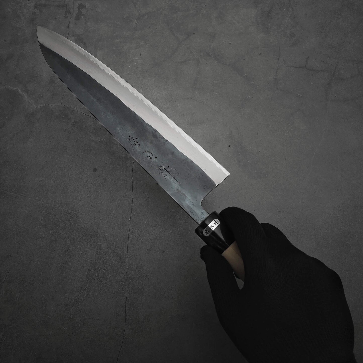 A gloved hand holds the Toyama kurouchi aogami#2 240mm gyuto by Toyama, featuring a sharp blade with engraved characters, set against a dark, textured background.