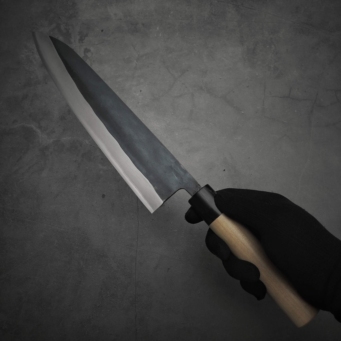 A hand in a black glove holds the Toyama kurouchi aogami#2 240mm gyuto—a gyuto with a black-finished aogami #2 blade and light wooden handle—against a dark, textured background.