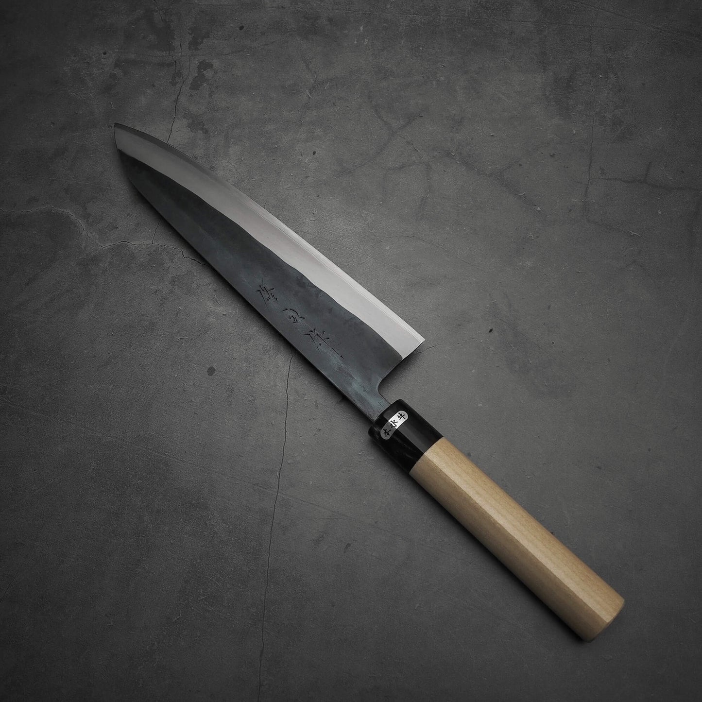 The Toyama kurouchi aogami#2 240mm gyuto, featuring a dark blade and light wooden handle, rests on a textured gray surface.