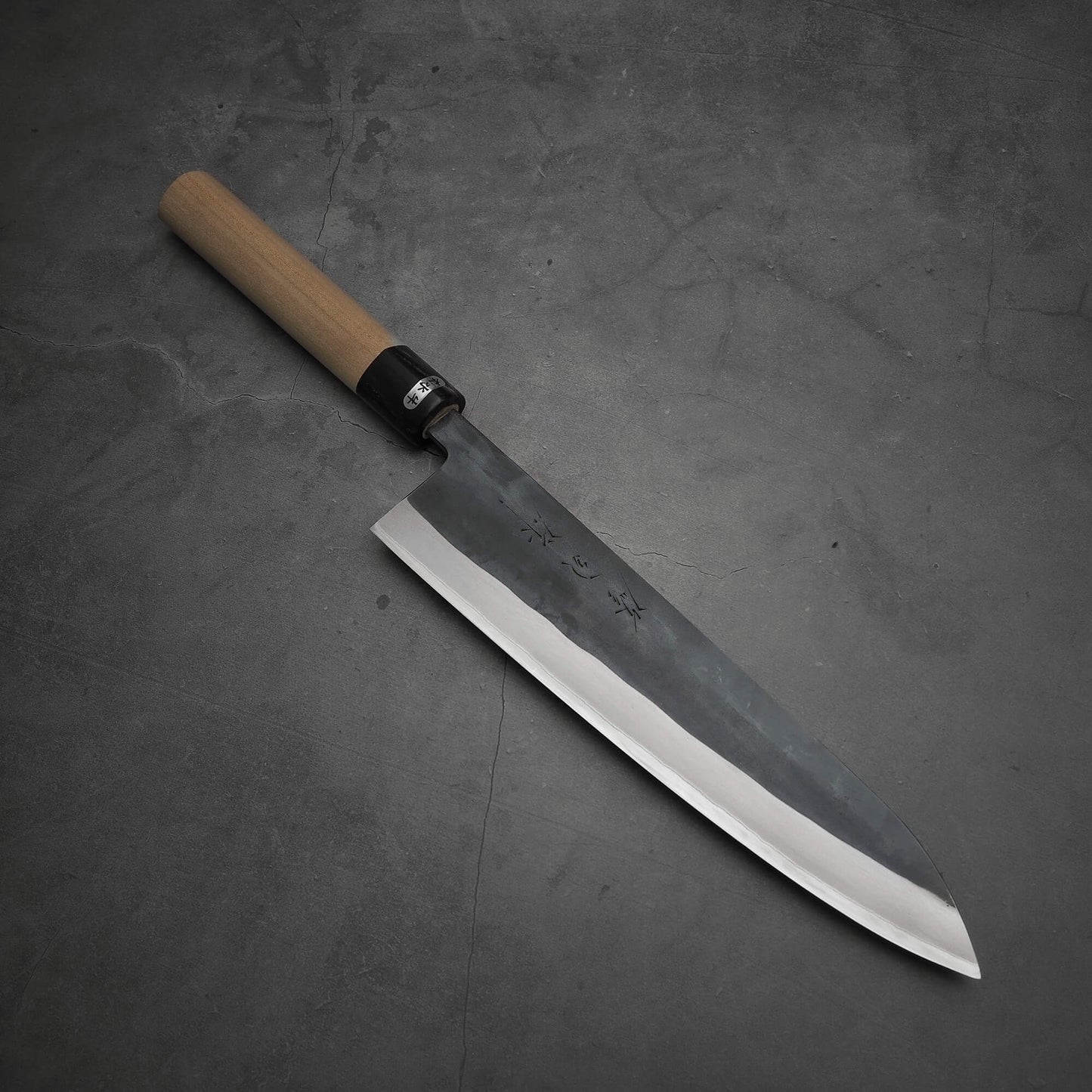 The Toyama kurouchi aogami#2 240mm gyuto, featuring a wooden handle and dark hammered blade, rests on a gray stone surface.