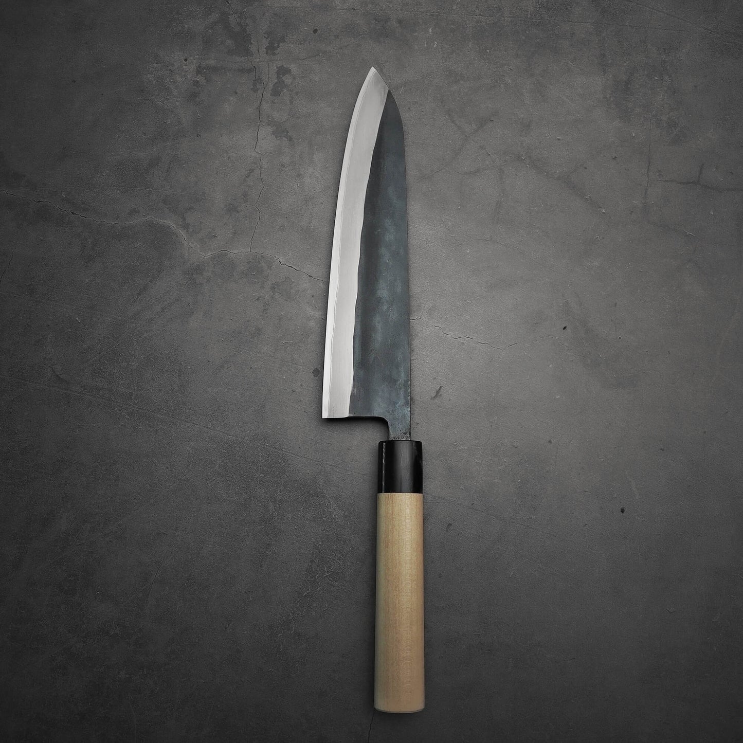 The Toyama kurouchi aogami#2 240mm gyuto, a Japanese-style chef’s knife from Toyama with a wooden handle and shiny pointed blade, rests on a dark gray textured surface.