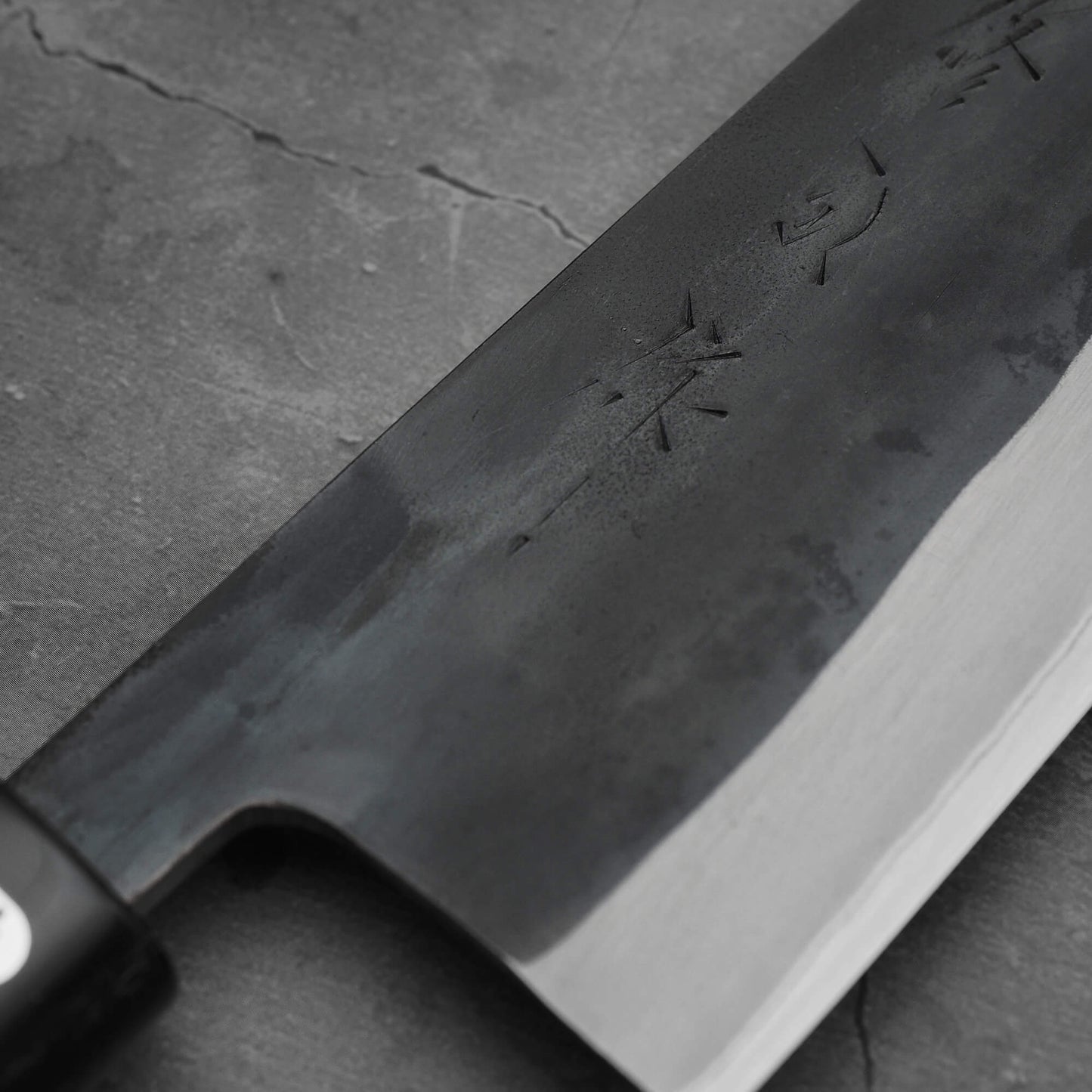 Close-up of the Toyama kurouchi aogami#2 240mm gyuto chef’s knife, featuring a matte dark blade with engraved characters, resting on a textured gray stone surface.