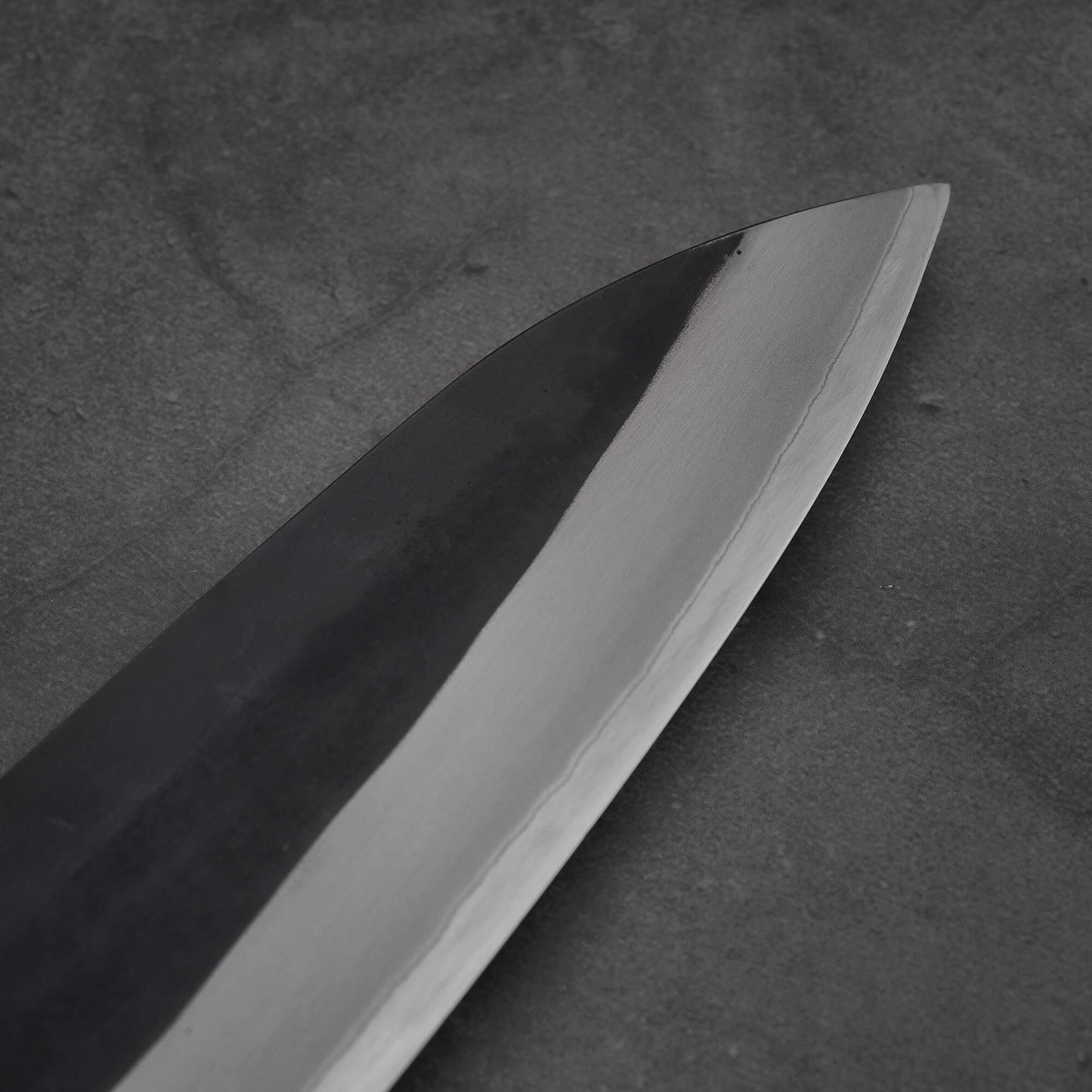 Close-up of the Toyama kurouchi aogami#2 240mm gyuto blade on a dark, textured surface. The sharp, shiny edge contrasts with the kurouchi finish and dark spine, showcasing Toyamas craftsmanship in aogami #2 steel.