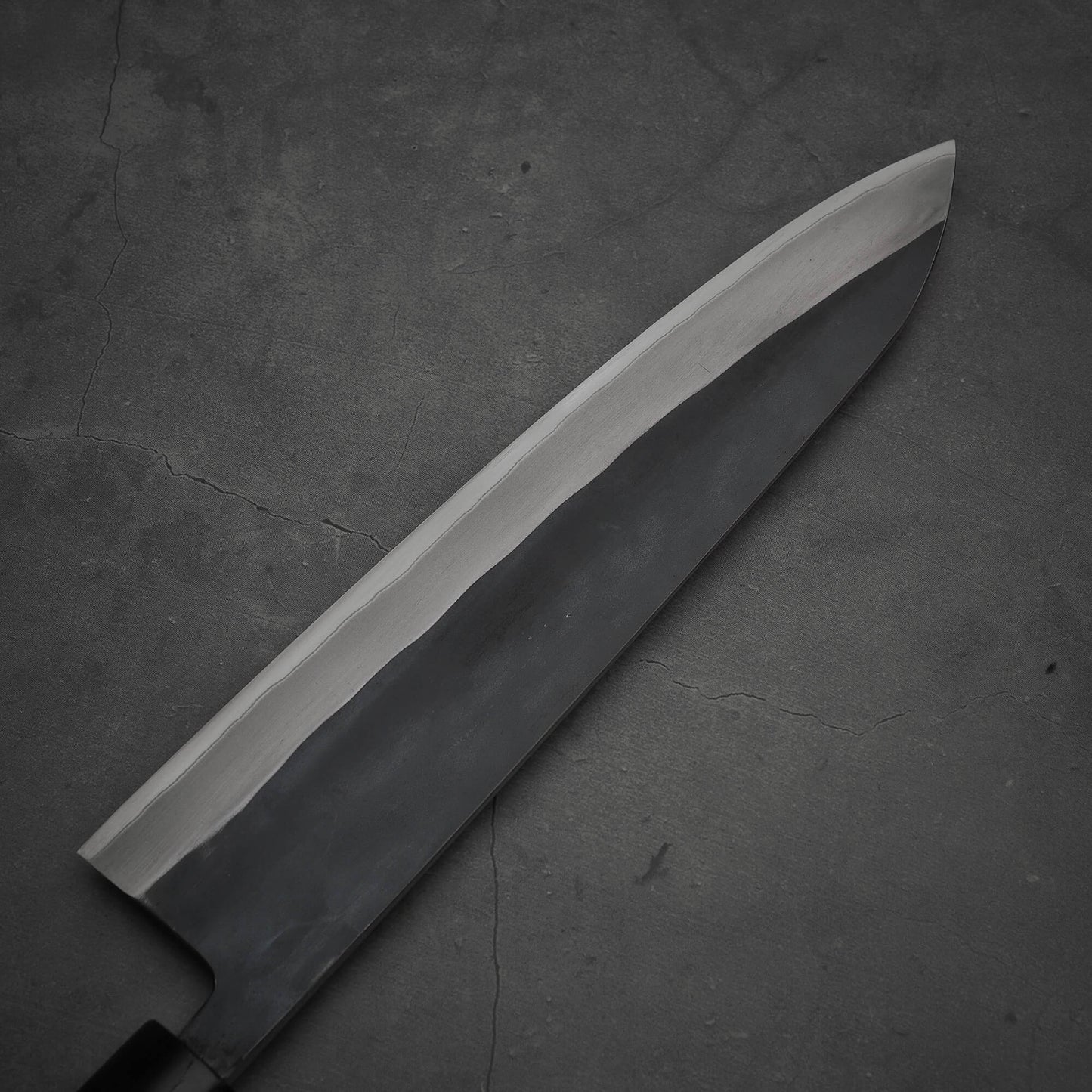 A close-up of the Toyama kurouchi aogami#2 240mm gyuto chef’s knife by Toyama, featuring a dark kurouchi blade with a lighter sharpened edge, positioned diagonally on a textured dark gray surface.