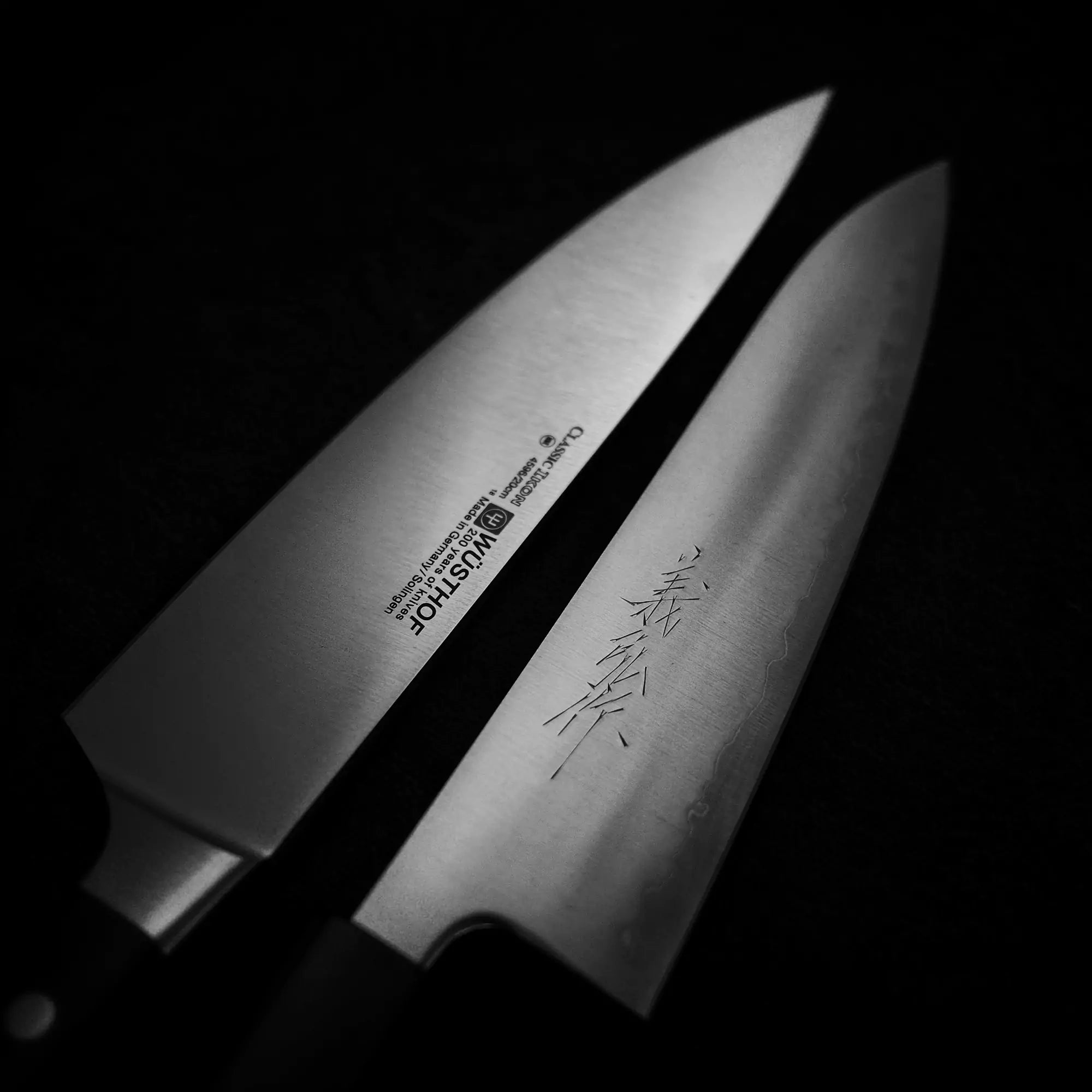Black and white close-up image of two chef's knives against a dark background. On the left is a Western chef's knife with inscriptions and branding, while on the right is a Japanese Gyuto chef's knife with distinct kanji characters etched on the blade.