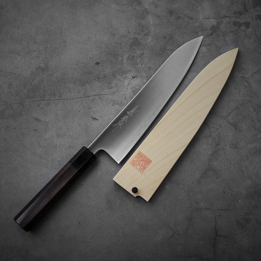 The Yoshihiro migaki AS gyuto 210mm kitchen knife, featuring a dark wooden handle and engraved blade, rests beside its light-colored wooden saya on a gray textured surface.