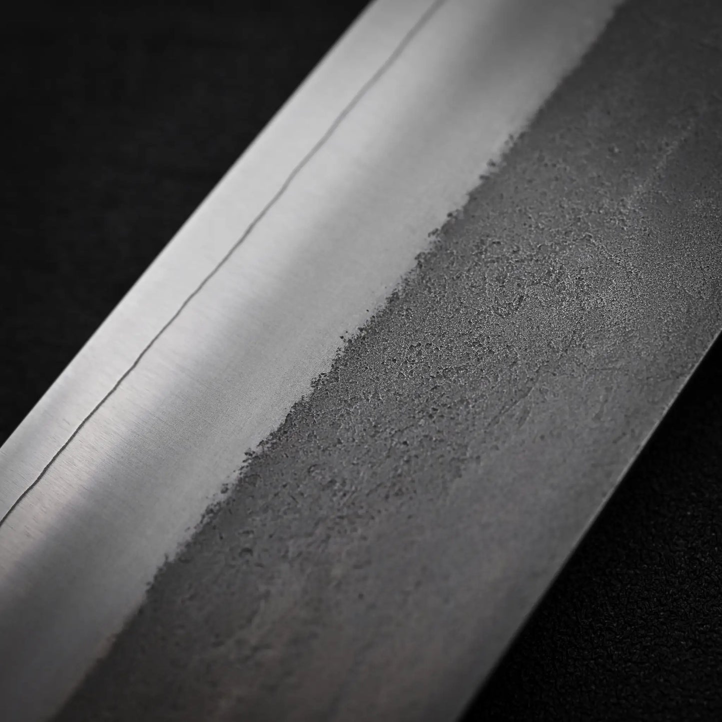 Close-up of the Yoshikane Hatsukokoro x Yoshikane nashiji SKD nakiri 165mm blade, highlighting its polished edge, wavy hamon line, and textured nashiji finish. The background is dark and blurred for emphasis.