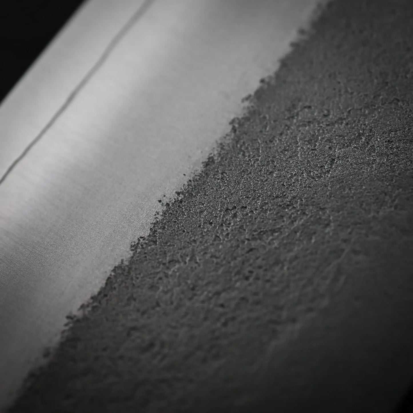A close-up of a surface showing a smooth, metallic left side and a rough, textured right—reflecting the unique nashiji finish seen on the Yoshikane Hatsukokoro x Yoshikane nashiji SKD nakiri 165mm knife.
