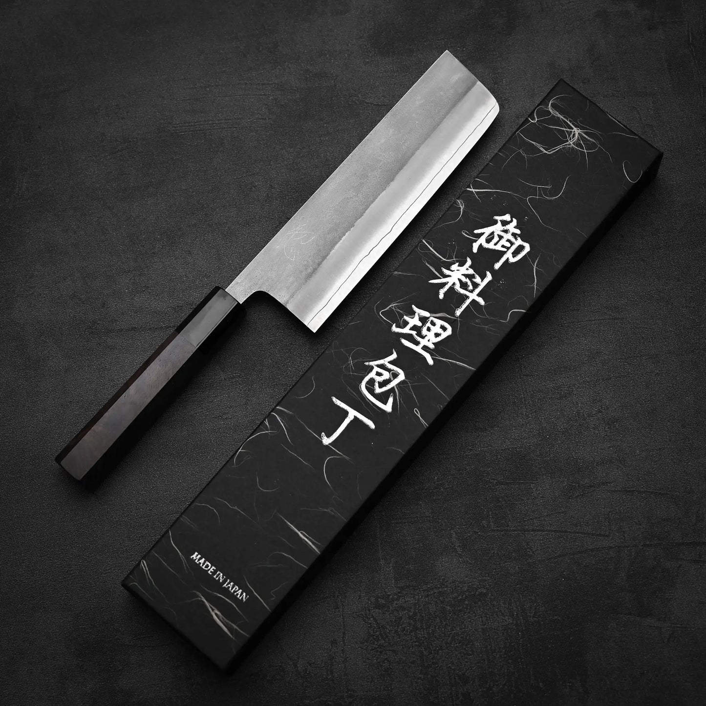A Hatsukokoro x Yoshikane nashiji SKD nakiri 165mm knife with a rectangular blade and dark handle rests beside a black box marked with white Japanese characters and MADE IN JAPAN on a dark surface. Brand: Yoshikane.