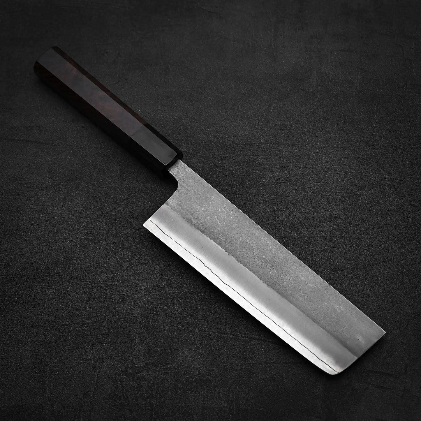 The Hatsukokoro x Yoshikane nashiji SKD nakiri 165mm knife by Yoshikane, featuring a rectangular blade and dark wooden handle, rests on a textured dark surface.