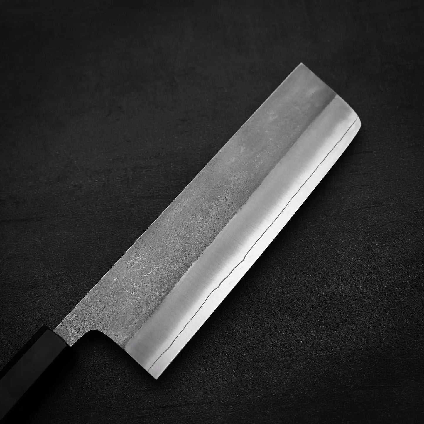 A Hatsukokoro x Yoshikane nashiji SKD nakiri 165mm Japanese knife by Yoshikane, featuring a rectangular blade and straight edge, lies on a dark textured surface. Its polished finish displays fine lines that showcase expert craftsmanship.