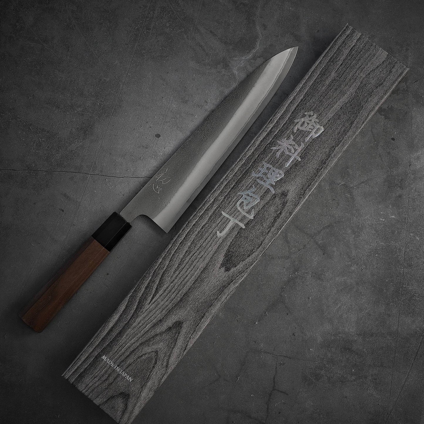 The Yoshikane nashiji shirogami#2 gyuto 240mm by Yoshikane, featuring a sharp blade and wooden handle, rests beside its engraved wooden case on a gray textured surface.