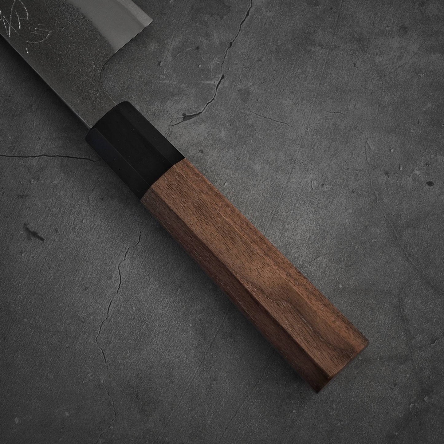 A close-up of the Yoshikane nashiji shirogami#2 gyuto 240mm knife with a wooden handle, showcasing part of the blade on a textured gray concrete surface.