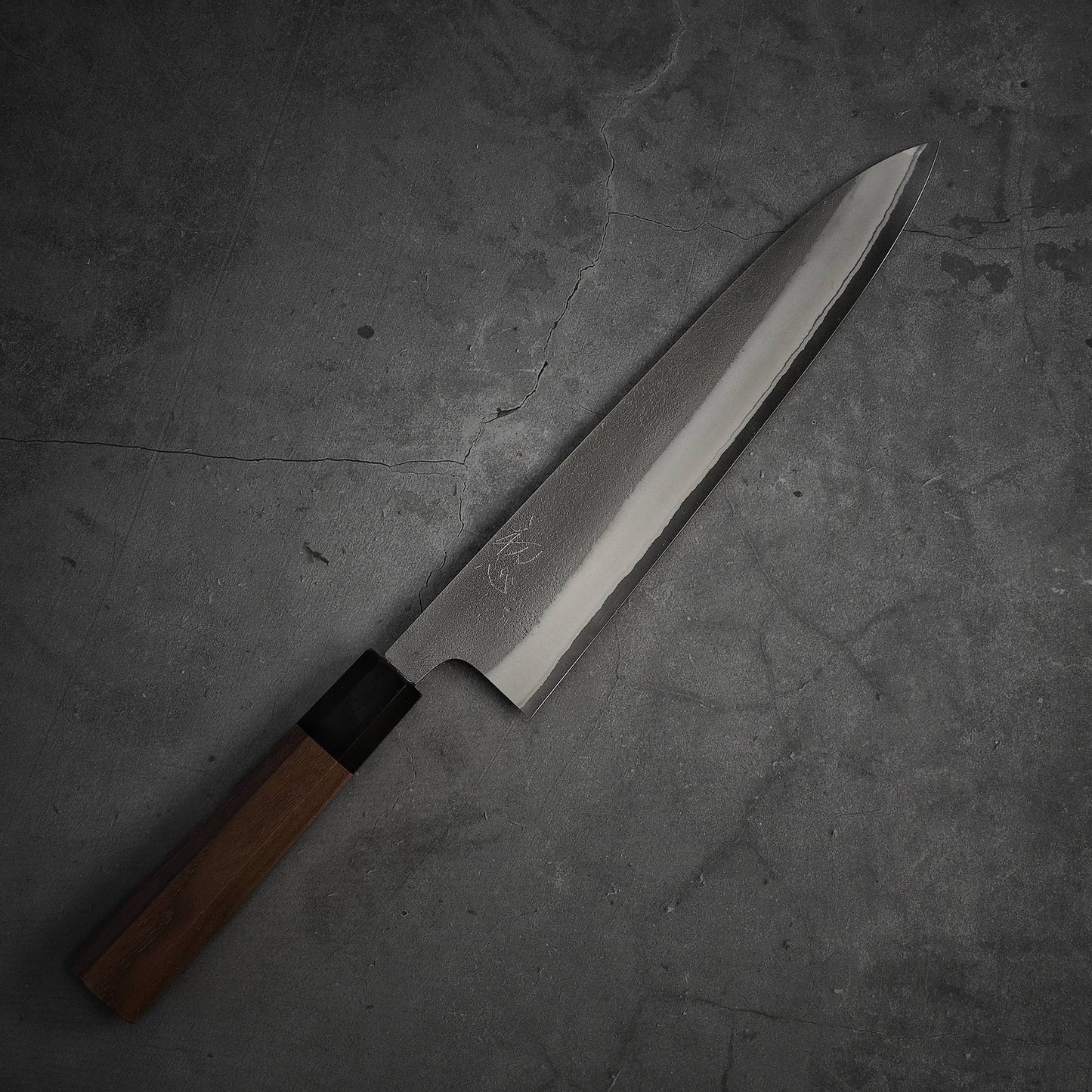 A Yoshikane nashiji shirogami#2 gyuto 240mm knife with a wooden handle lies on a gray, textured concrete surface. Its shirogami#2 steel blade, by Yoshikane, has Japanese characters etched near the handle.