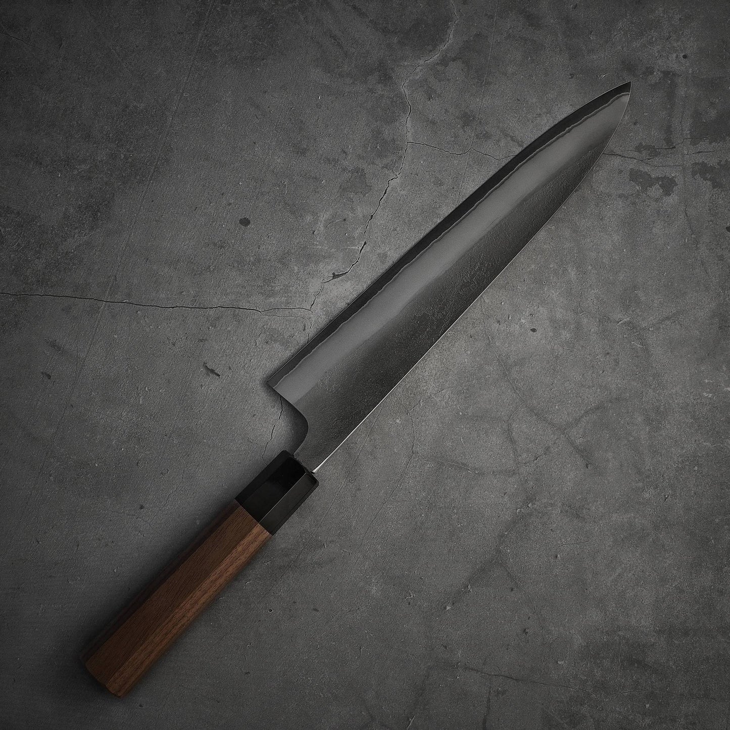 The Yoshikane nashiji shirogami#2 gyuto 240mm, featuring a dark wooden handle and a sharp, shiny blade, is displayed diagonally on a textured gray stone surface.